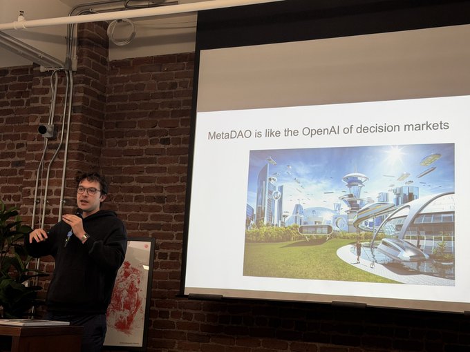 A man in a black hoodie giving a presentation in front of a brick wall, with a projected slide reading 'MetaDAO is like the OpenAI of decision markets' accompanied by a futuristic cityscape illustration.