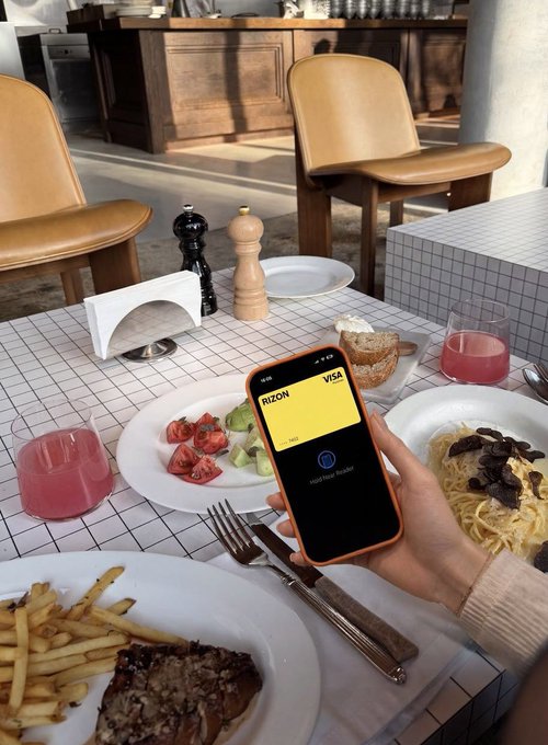 A person holds up a smartphone displaying the "RIZON" Visa card app over a restaurant table with meals and pink drinks, promoting Rizon's new physical crypto card with the first 1,000 available free.