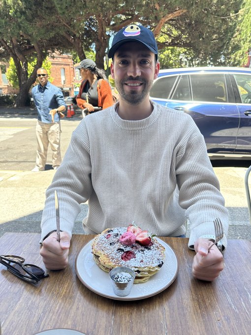 The image features a man sitting at an outdoor table with a plate of pancakes topped with strawberries and whipped cream. He is wearing a baseball cap and has his hands around a fork, ready to enjoy his meal. In the background, there are two other people present - one person standing behind the man and another person walking by on the sidewalk. The scene suggests that this could be a casual dining experience or an outdoor gathering where friends have come together for breakfast.
