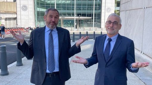Two men in business suits stand outside a modern glass building, gesturing with open palms, likely illustrating Ripple's Chief Legal Officer Stuart Alderoty's call for senators to hear directly from the 67 million American crypto holders rather than industry lobbyists before voting on the CLARITY Act.