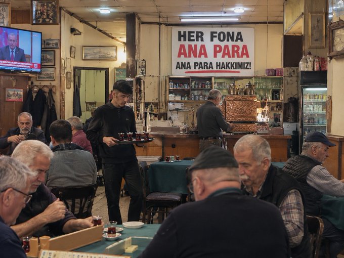 A crowded Turkish café interior with men sitting at tables playing games; a sign reads 'HER FONA ANA PARA ANA PARA HAKKIMIZ!' and a waiter carries a tray of tea.