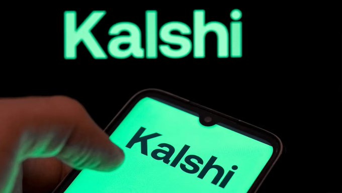 A close-up of a smartphone displaying the 'Kalshi' logo in green, held in front of a black background with a larger 'Kalshi' logo illuminated behind it.
