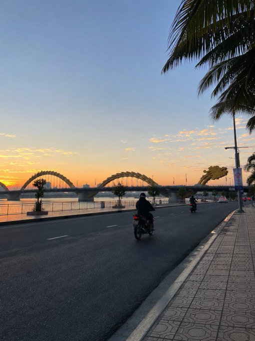 A motorcyclist rides along a riverside road at sunset, with the iconic golden Dragon Bridge silhouetted against the warm orange sky.

The image perfectly captures the "beautiful note" mentioned in the post—the serene sunset scene and elegant bridge embody the peaceful, hopeful close to the week, while the lone rider heading toward the horizon mirrors the reflective mood of wrapping up Friday.