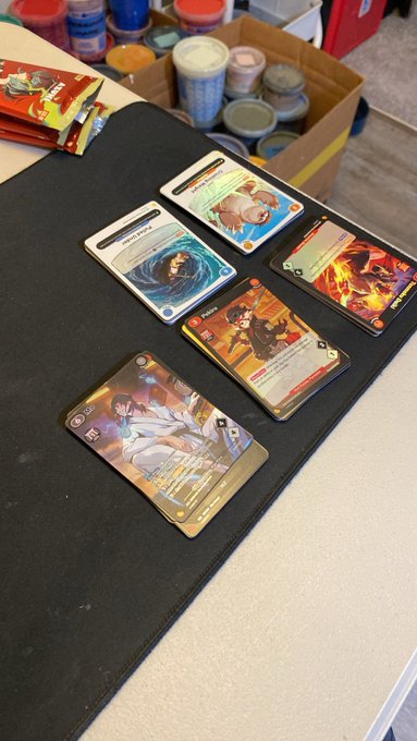 Several trading cards featuring anime-style character artwork are arranged on a black mat on a table, with a box of card supplies visible in the background.