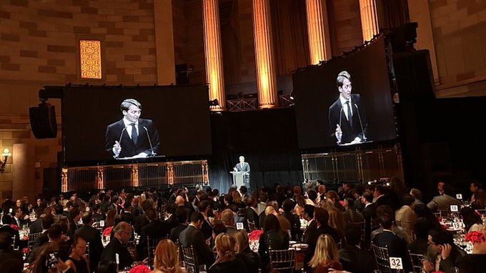 A formal gala event features a speaker at a podium broadcasting on two large screens to a seated audience, with the screens showing the speaker wearing glasses and a suit.