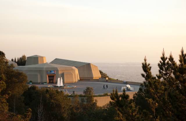 A modern building with angular architecture sits on a hilltop overlooking a hazy city at sunset, with pine trees in the foreground.