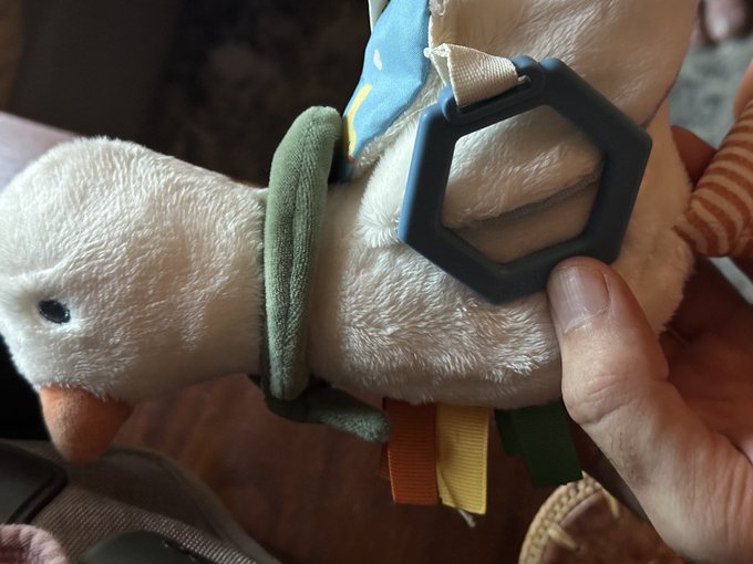 A close-up of a hand holding a beige plush bird toy equipped with a black plastic D-ring clip and a blue patterned strap, featuring colorful ribbon tags on its underside.