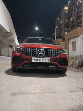 The image features an orange Mercedes-Benz car parked outside of a building at night, with its headlights turned on. The car has a license plate that reads "MHS 1313". In the background, there are several buildings visible behind the car. The text in the image suggests that someone is trying to sell the car for $BTC or Bitcoin, and it seems like an advertisement for a cryptocurrency exchange.