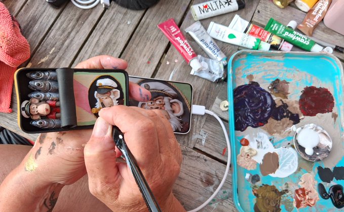 An artist meticulously hand-paints a "Curtis" Bored Ape character on a phone case, referencing the original print by @_Solvex_, @BoringBrush, and @BoredApeYC visible on a second phone beside them, with acrylic paints and brushes scattered on the wooden surface.