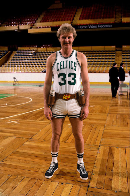 A vintage photograph of NBA player Larry Bird wearing a white Celtics jersey with the number 33, standing on a basketball court with water bottles tucked into his shorts, in front of empty arena seats.