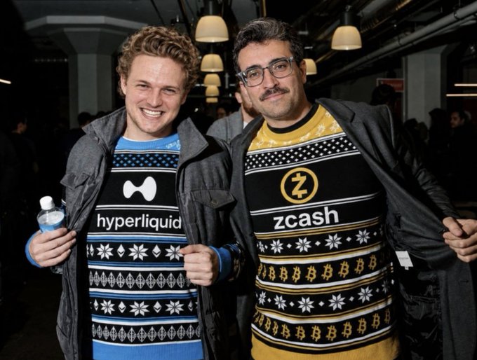Two men smiling and holding open their jackets to show festive holiday sweaters; the left one wears a blue sweater with the "hyperliquid" logo and the right one wears a yellow and black sweater with the "zcash" logo.
