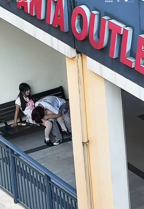 Under a red sign partially reading 'ANIA OUTLE', two young women in matching outfits sit on a black bench, with one leaning her head on the other's shoulder.