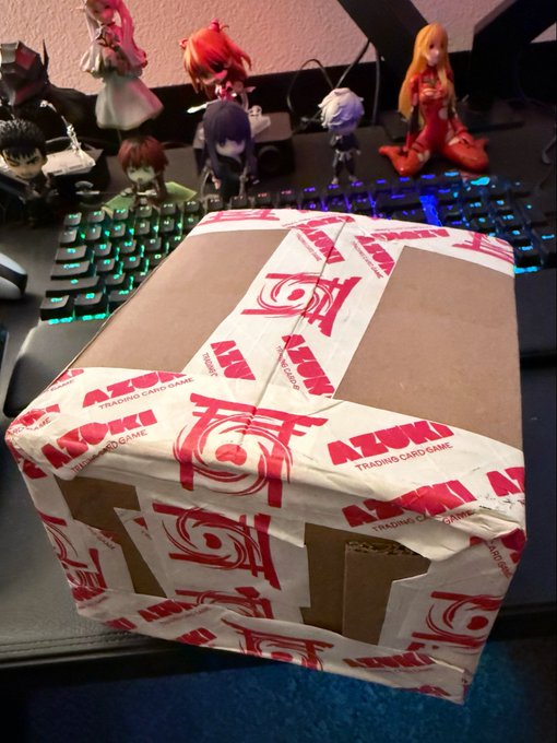 The image features a cardboard box with red and white stripes, sitting on top of a desk next to a keyboard. The box has been wrapped in red paper and has a sticker that reads "Booster Box Round Deux Ya’ll thinking what I'm thinking?". This suggests that the box might be a gift or a surprise for someone, possibly related to anime or gaming.