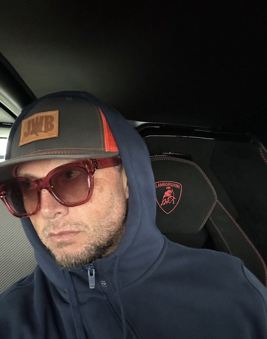 The "wen lambo?" caption is answered here—a man sits inside a Lamborghini, wearing a hoodie with red-tinted sunglasses and a cap featuring an "LAMB" leather patch, with the Lamborghini bull logo embroidered on the seat behind him.