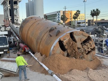 Construction workers operate a large cylindrical drill at an urban construction site, with dirt piled up and city buildings and palm trees in the background.