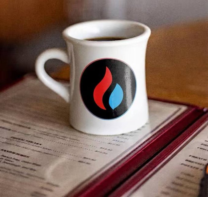 A white ceramic mug filled with dark coffee sits on an open book, featuring a circular logo with red and blue flame-like shapes on a black background.