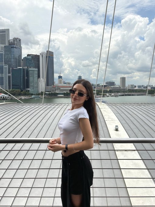 A woman wearing sunglasses, a white crop top, and black skirt poses on a rooftop overlooking a city skyline and waterfront.