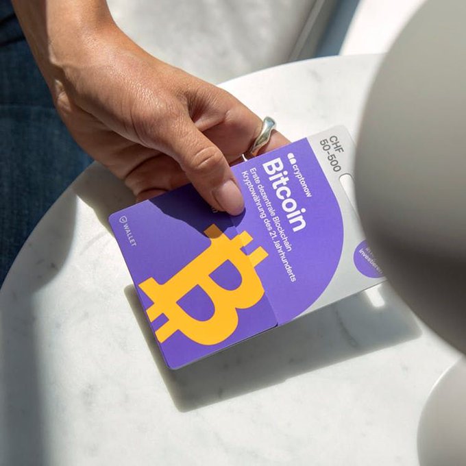 A person holds a purple "Bitcoin Starter Kit" gift card from Swiss company Cryptonow, displaying the iconic orange Bitcoin "₿" symbol and denominations ranging from CHF 50-500, demonstrating the new in-store crypto purchasing option now available at French retailers like Carrefour.