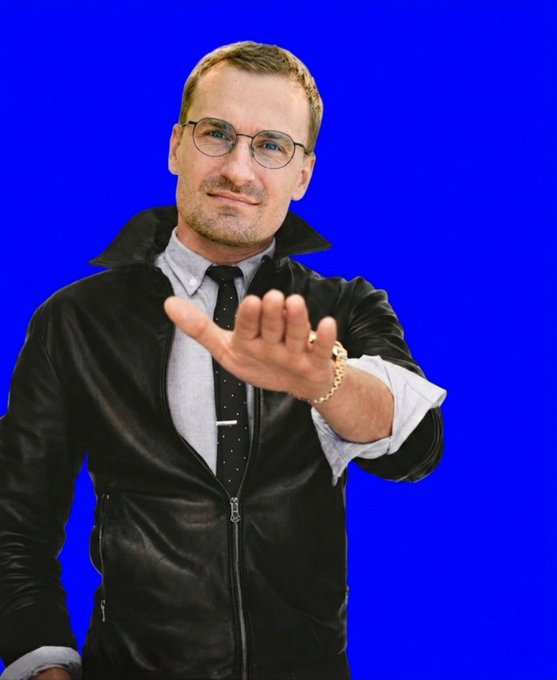 A man with glasses and a black leather jacket extends his hand forward against a solid blue background.