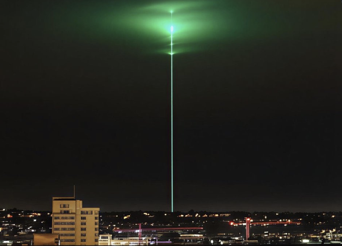 A nighttime cityscape featuring a striking green laser beam shooting vertically into the sky from the urban horizon.
