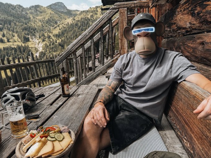 A person wearing a BAPE monkey mask and cap relaxing on a wooden deck overlooking mountains, with a beer, helmet, and charcuterie board on the table.