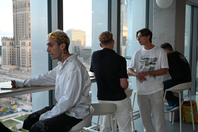 Several young people with bleached blonde hair stand and sit in a high-rise space with floor-to-ceiling windows overlooking a city; one wears a white t-shirt with a Calvin Klein logo and another wears a black hoodie reading "blcg".