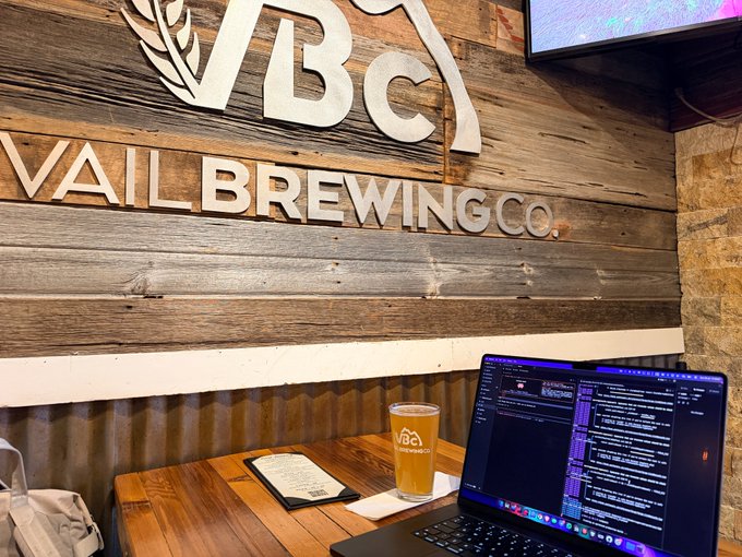 A laptop displaying code is set up at a wooden table inside Vail Brewing Co., accompanied by a glass of beer with the brewery's logo, visually representing the intersection of technology and craft brewing highlighted in the post.