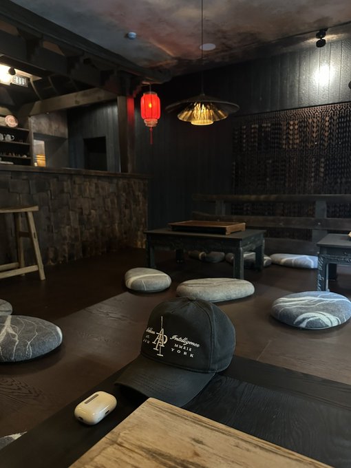 A black cap reading "Atrium Plaza House New York" rests on a wooden table in a dimly lit lounge featuring Asian-inspired decor, red lantern, and floor cushions, illustrating the post's point about how branded merchandise sparks conversations and shares crypto project stories organically.