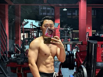 A mirror selfie of a muscular, shirtless Asian man taking a photo with his smartphone in a weight room with red equipment in the background.