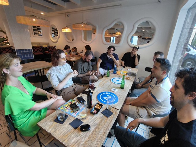 The image shows a group of people sitting around a dining table, engaged in conversation. There are at least nine visible individuals, with some seated on chairs while others stand or sit on the floor. The table is set with various items such as wine glasses, cups, and bottles, suggesting that they might be enjoying drinks together. A few cell phones can also be seen on the table.

The room has a white wall behind them, which features several mirrors arranged in an interesting pattern. There are two chairs visible near one of the mirrors, possibly indicating a more formal or upscale setting for their gathering. The overall atmosphere appears to be relaxed and social as they share stories and enjoy each other's company.