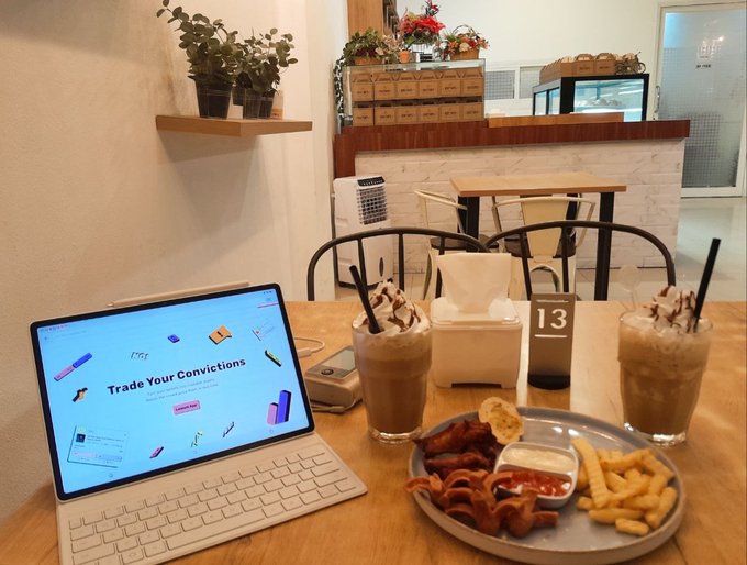 A laptop displays a Coinbase webpage reading "Trade Your Convictions" alongside two iced coffee drinks and a snack platter of fries, onion rings, and dipping sauces on a wooden table in a café setting, fittingly tying the "Trade Your Convictions" slogan to the "cooking" market theme.