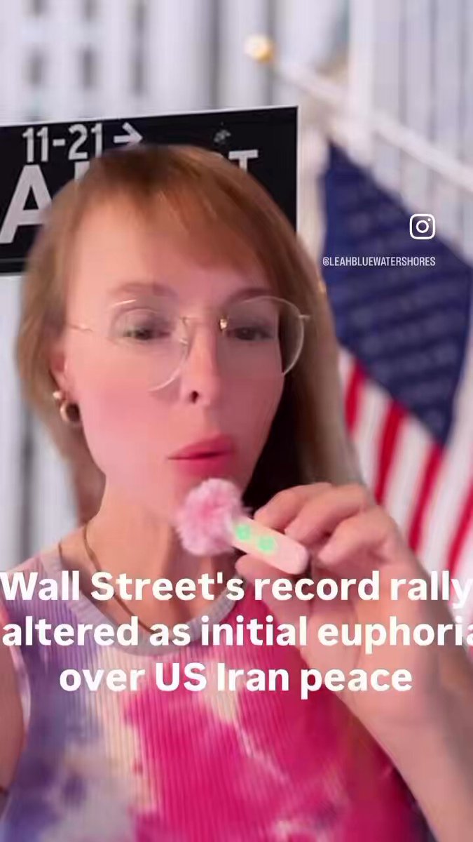 The image features a woman wearing glasses, holding a pink flower-shaped object (likely a lollipop) in her mouth while standing next to a sign that says "Wall Street's record rally altered as initial euphoric over US Iran peace". The text on the sign suggests that there is an ongoing rally at Wall Street involving the alteration of initial euphoria towards a more aggressive stance. This could be related to the tensions between the United States and Iran, with the US imposing economic sanctions against Iran in exchange for certain concessions from Iran.
