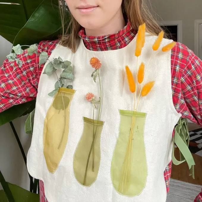 A person wearing a red plaid shirt under a white vest that has three clear plastic pouches serving as vases for eucalyptus, orange flowers, and dried wheat.
