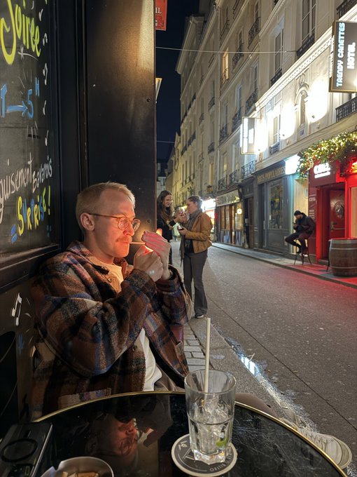 Evening street scene in what appears to be a European city, with a young man in a plaid jacket and glasses lighting a cigarette at a bar, while other patrons converse on the sidewalk near a cigar lounge hotel.