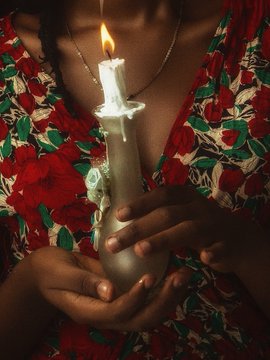 A pair of hands gently cradles a lit white candle, with the flame illuminating a red floral-patterned garment in a dimly lit setting.
