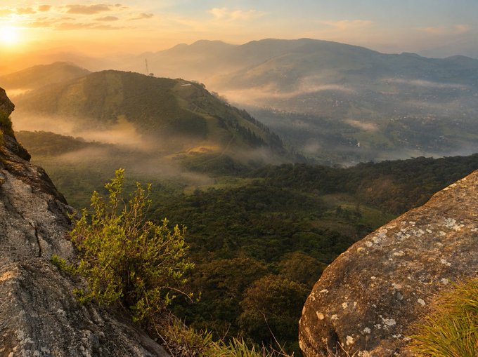 A scenic mountain landscape at sunrise with mist rolling over forested hills, viewed from between two large rock formations.