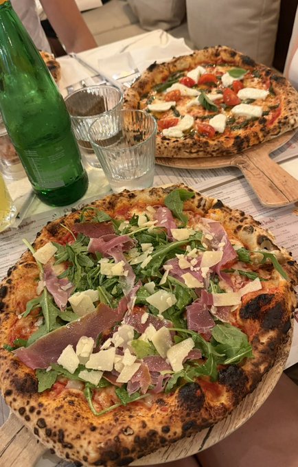 Two wood-fired Neapolitan-style pizzas are served on wooden boards at a restaurant table—one topped with prosciutto, arugula, and shaved parmesan, the other featuring fresh mozzarella, tomatoes, and basil—a delicious-looking "smash" choice for the pizza lovers.