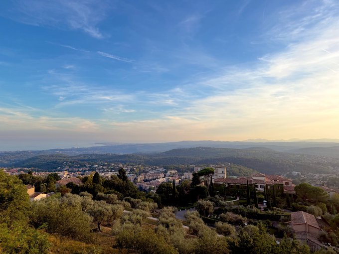 A scenic elevated view overlooking a Mediterranean landscape with rolling hills, a coastal town, the sea in the distance, and a villa with olive trees in the foreground under a partly cloudy sky at sunset.