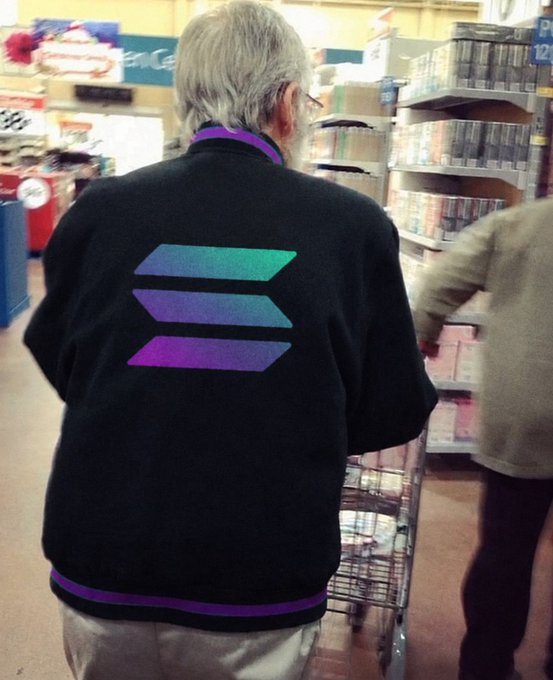 An elderly person with gray hair stands in a grocery store aisle wearing a black jacket featuring the three-bar Solana logo in purple and teal, while pushing a shopping cart.