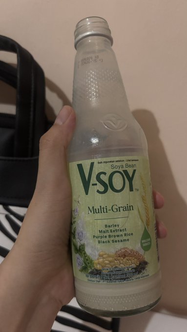 A hand holds up a glass bottle of V-Soy Multi-Grain soya bean milk, listing ingredients including barley, malt extract, purple brown rice, and black sesame.
