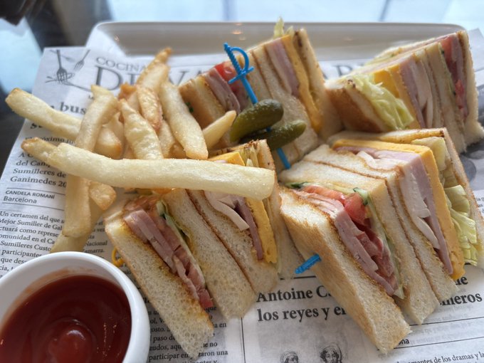 A club sandwich cut into quarters with ham, turkey, cheese, lettuce, and tomato, served with french fries, pickles, and a small cup of ketchup on a tray lined with newspaper-style paper.