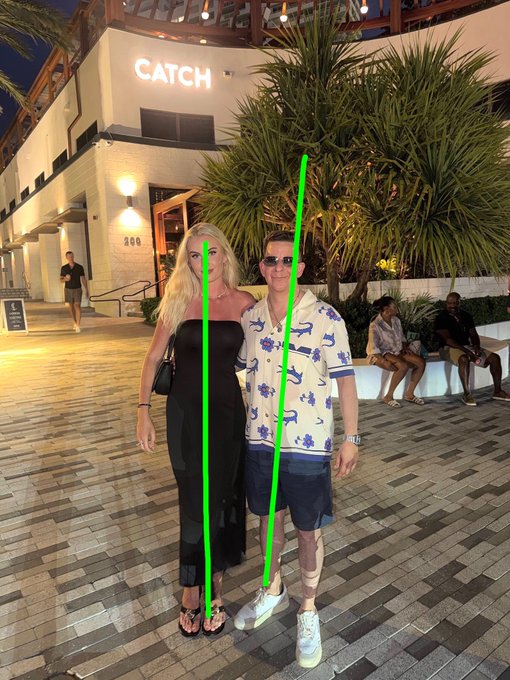 A couple poses outside a restaurant called CATCH, with green lines drawn to highlight the height difference between the tall woman and the shorter man.