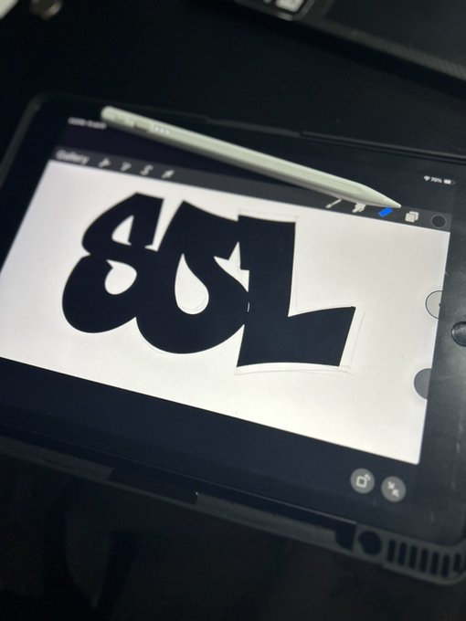 A photo of an iPad on a dark surface with a white stylus resting on it, displaying the Procreate app where someone is drawing bold black graffiti-style lettering on a white canvas.