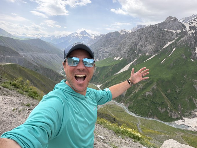 A man wearing sunglasses and a teal hoodie takes an excited selfie with arms outstretched against a stunning mountain valley landscape with snow-capped peaks.