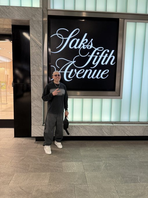 A bald man wearing sunglasses, a black shirt, baggy gray pants, and white sneakers poses with a peace sign in front of the illuminated 'Saks Fifth Avenue' storefront sign.
