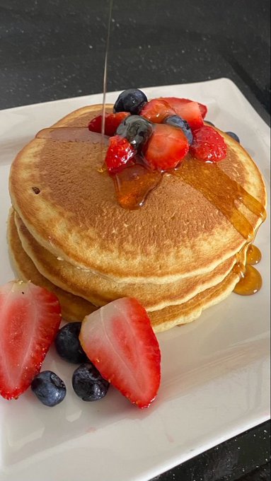 A stack of pancakes topped with fresh blueberries and raspberries on a white plate, with syrup being drizzled over the top.