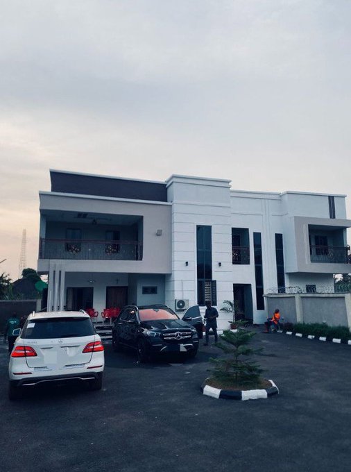 A modern white two-story house with a paved parking area featuring a white Mercedes SUV, a black Mercedes SUV, and a red car, with a few people visible near the entrance.