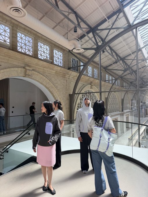 A group of young people wearing matching shirts that say 'Start Something' stand in a modern, sunlit indoor space with large arched windows and exposed steel beams.