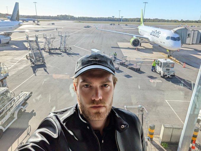 A bearded man in a black leather jacket and cap takes a selfie inside an airport terminal, with a parked "Bamboo" airline plane visible on the tarmac behind him and ground crew working nearby. The image captures the moment after his delayed airport security experience mentioned in the post, with his nickname "Rocky" referenced alongside the Bitcoin symbol.
