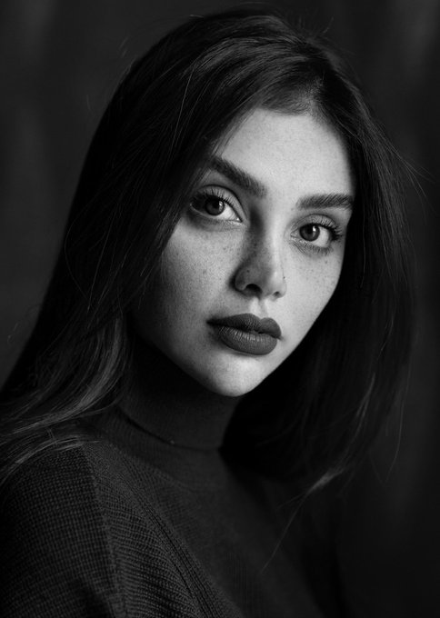 A black and white portrait photograph of a young woman with long dark hair and freckles, wearing dark lipstick and a turtleneck sweater, gazing at the camera.